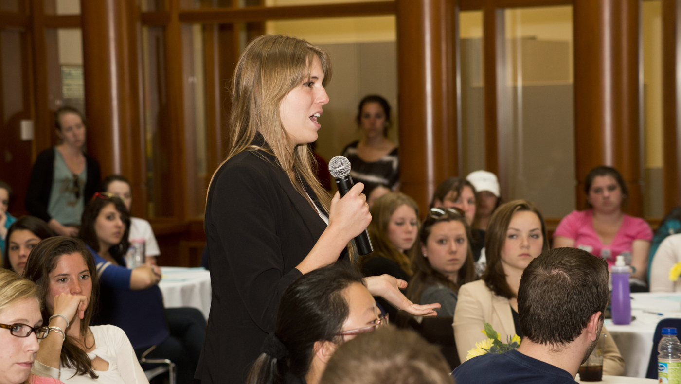 A graduate student with a microphone asking a question during a program as others listen.